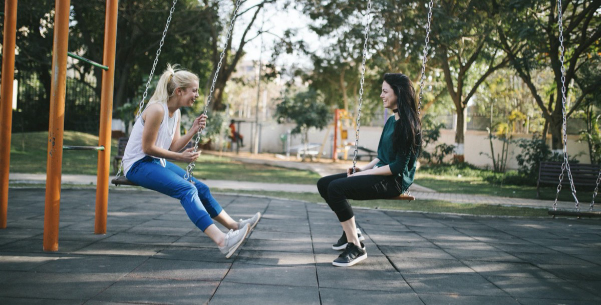 Two girls sitting on a swing set talking and happy. 
