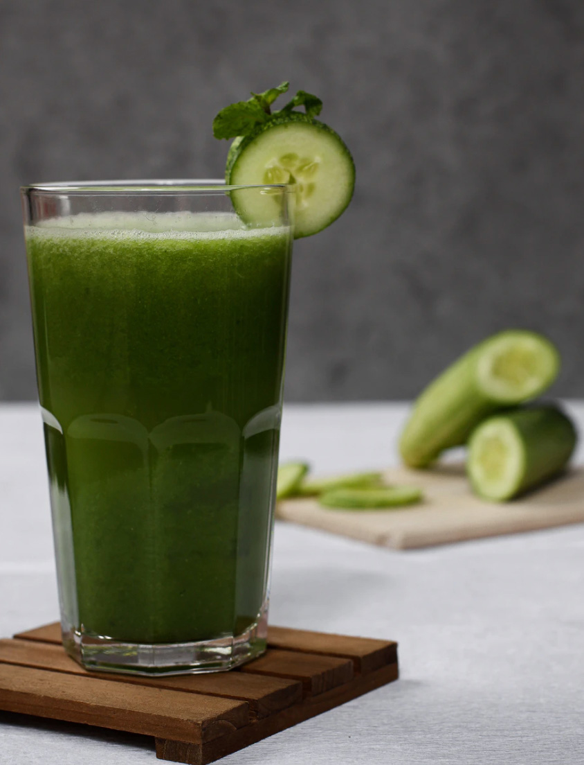 A cup of green juice with cucumbers in the background 